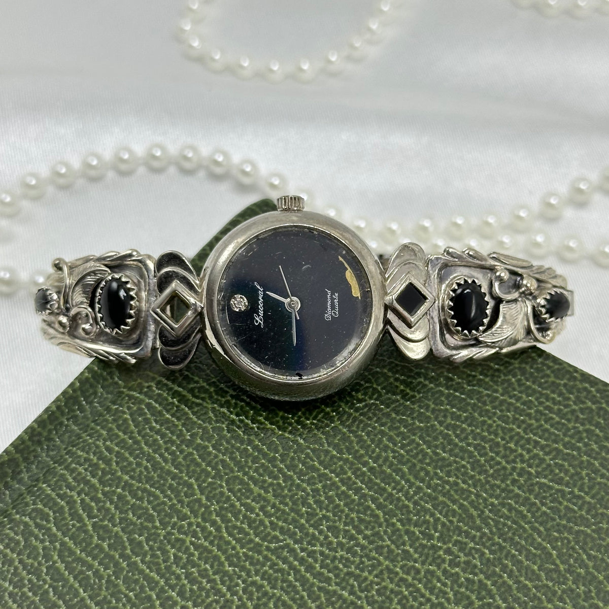 Vintage Navajo-style Lucoral diamond quartz watch with round black dial, sterling silver leaf and scrollwork watch tips laid with polished black onyx stones on a stretchable band. Missing one onyx stone.&nbsp;