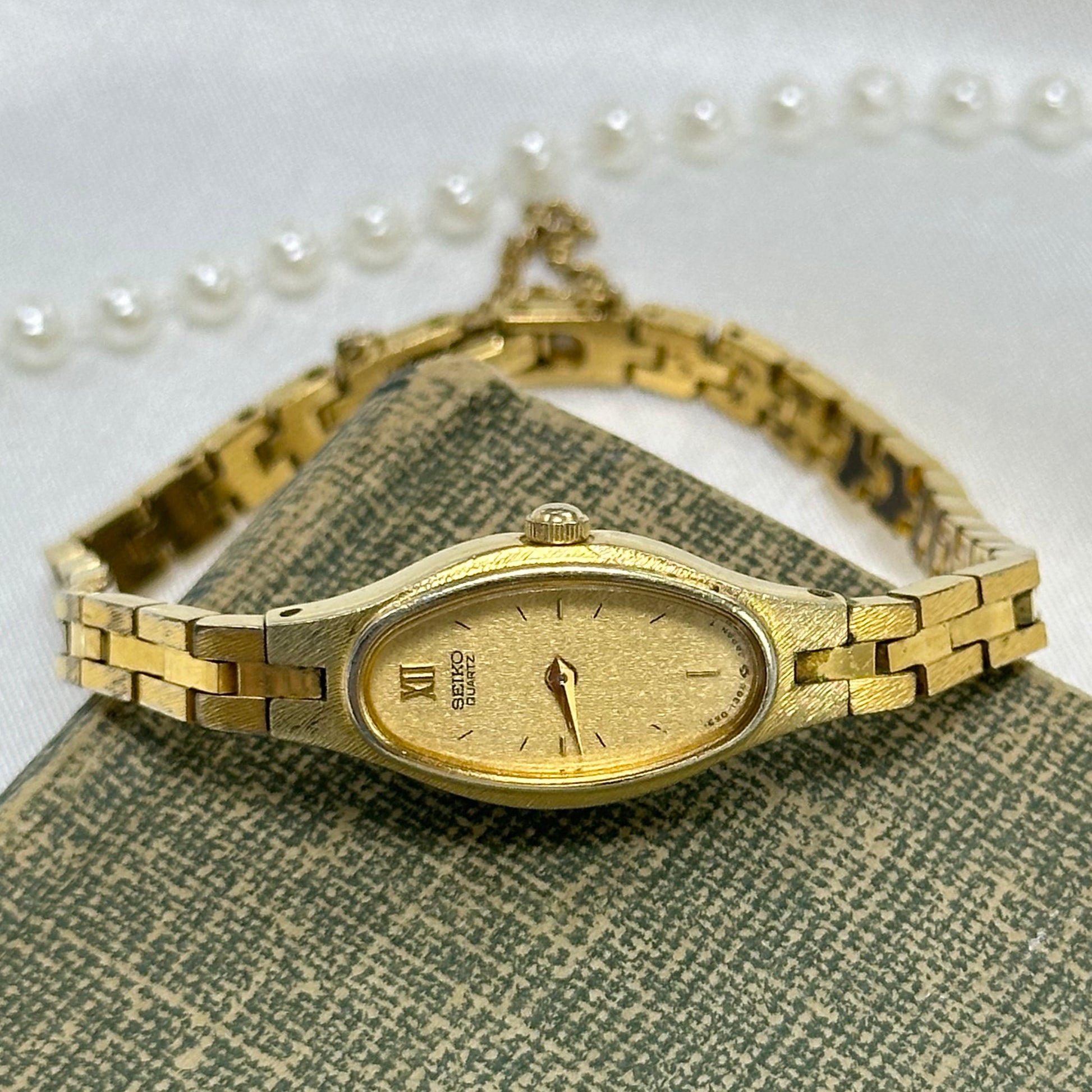 Dainty gold tone oval dial Seiko with slender clasp link band and safety chain.