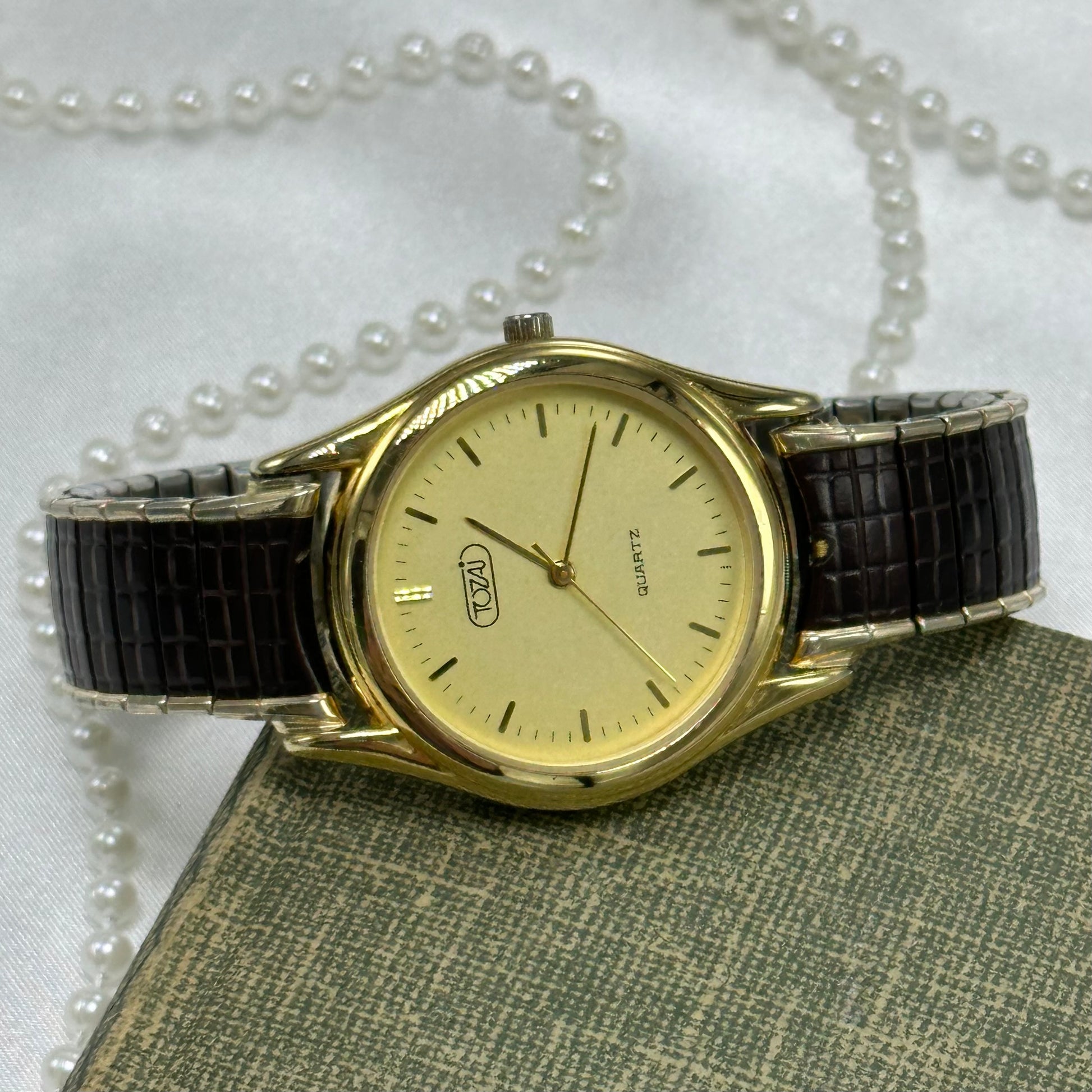 Gold tone vintage round dial Tozai watch with brown stretchable band and quartz movement.