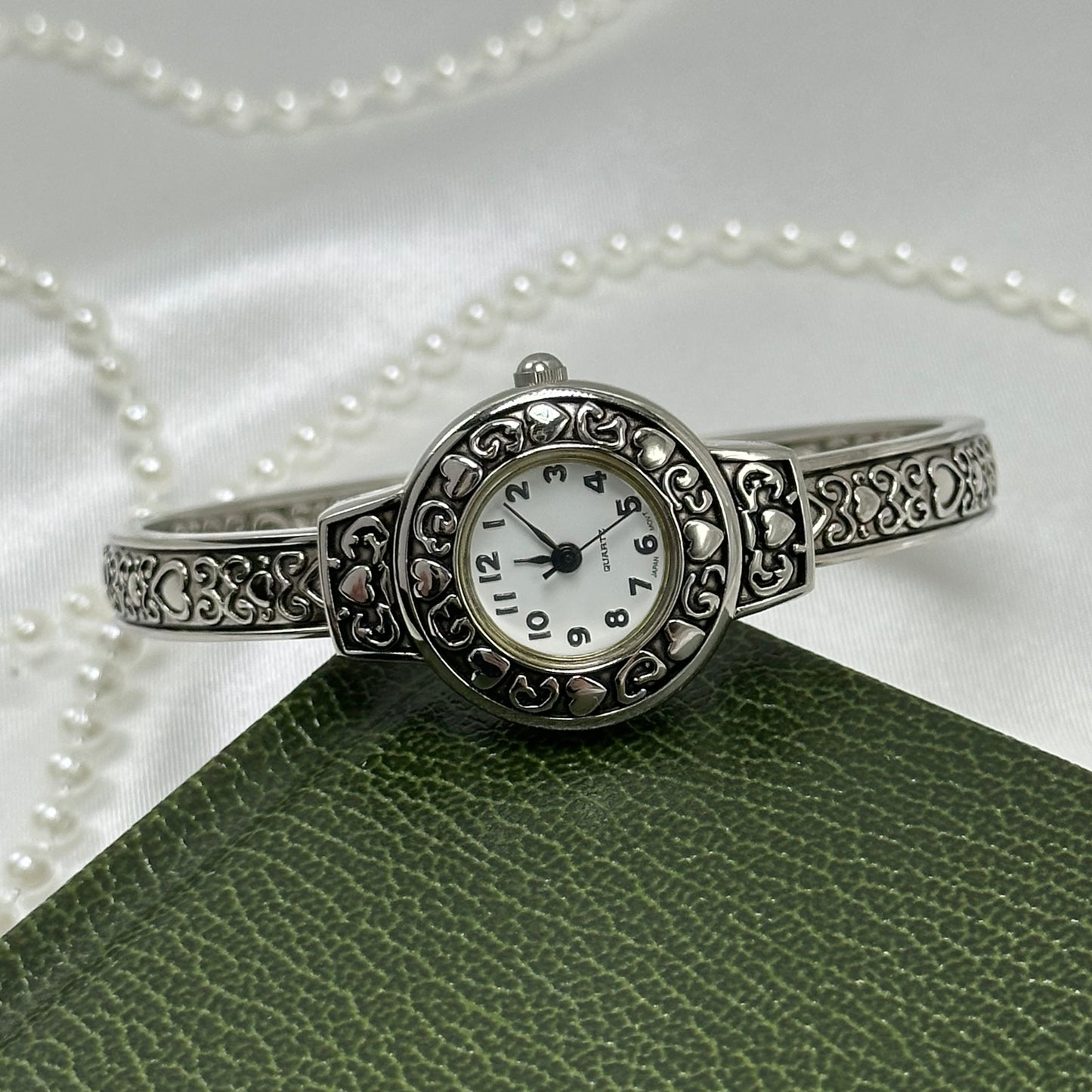 Silver tone cuff watch with round dial and detailed heart design.