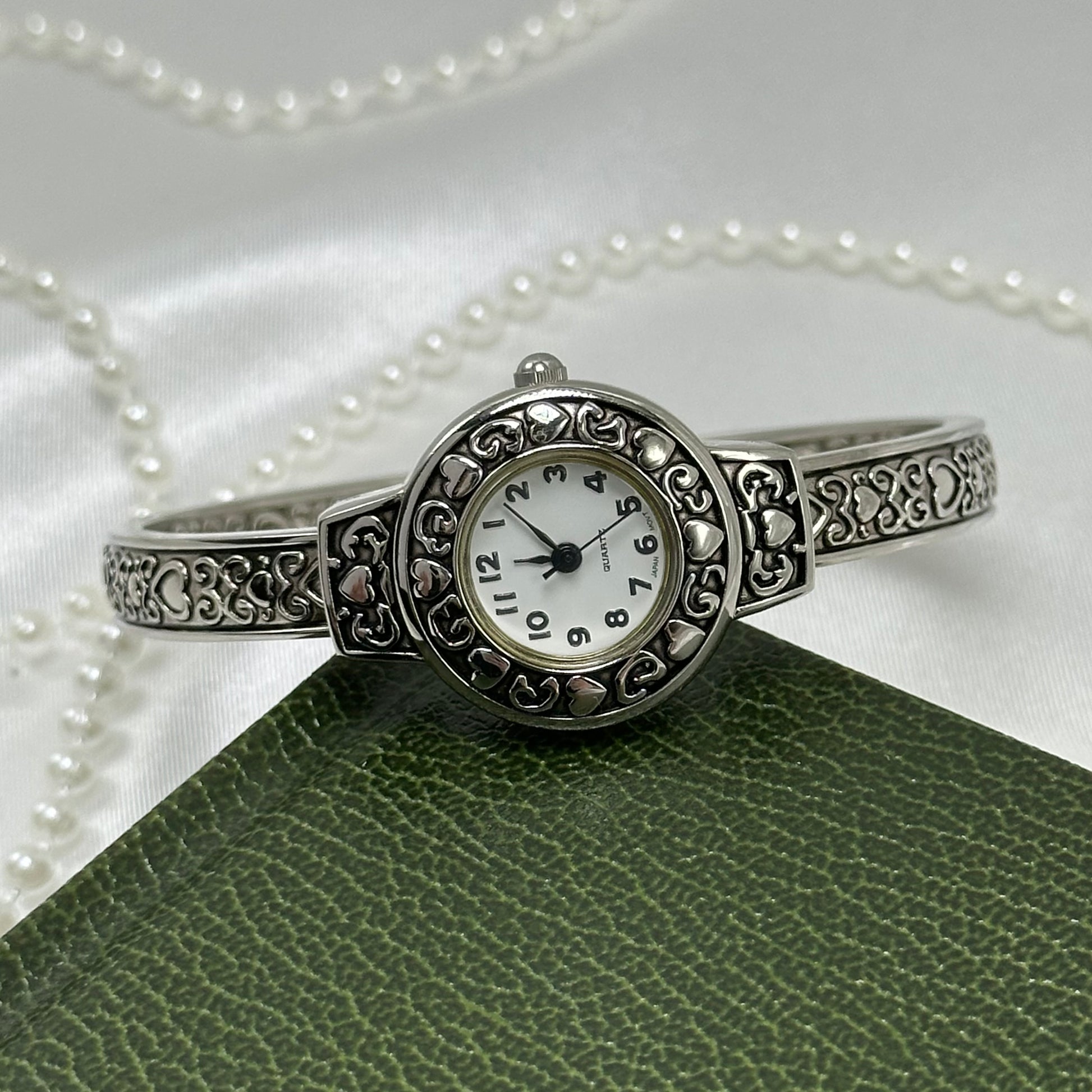 Silver tone cuff watch with round dial and detailed heart design.