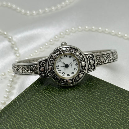 Silver tone cuff watch with round dial and detailed heart design.