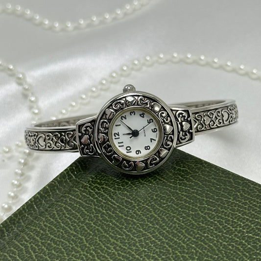 Silver tone cuff watch with round dial and detailed heart design.