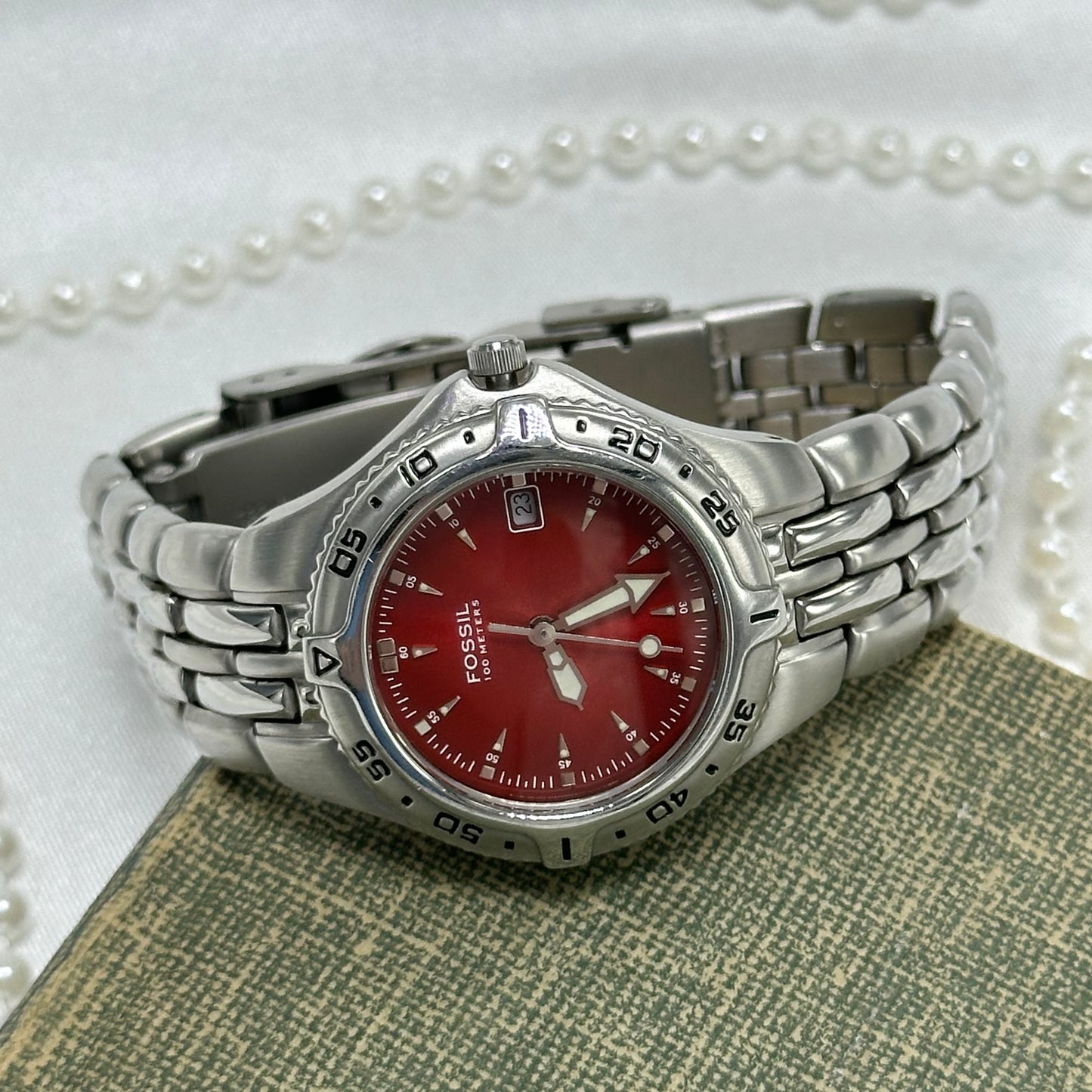 Silver tone red dial Fossil watch with double locking fold over clasp and date display. Hands are luminous. Numbers around the dial spin. Water resistant up to 330 ft (100 meters).