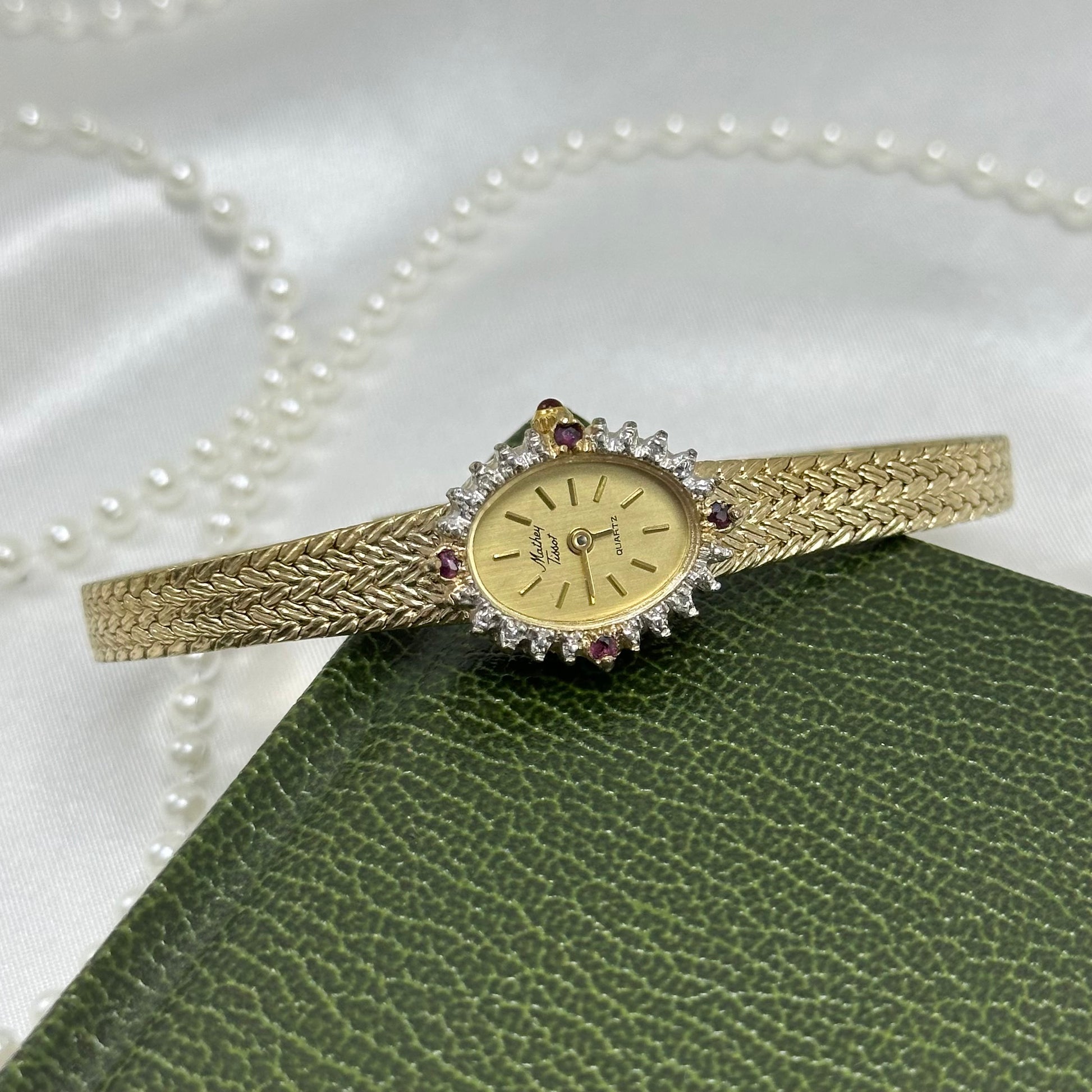 Vintage gold tone Mathey Tissot watch with lab grown diamonds and rubies around a dainty oval dial, slender metal adjustable band, and red jewel crown.
