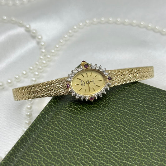 Vintage gold tone Mathey Tissot watch with lab grown diamonds and rubies around a dainty oval dial, slender metal adjustable band, and red jewel crown.