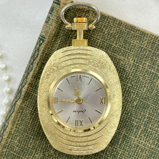 Vintage gold tone manual winding Endura Swiss made pocket watch with red jewel crown and roman numeral dial.