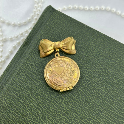 Vintage gold tone round locket with engraved heart design attached to a bow pin. Has photos inside that can be removed.