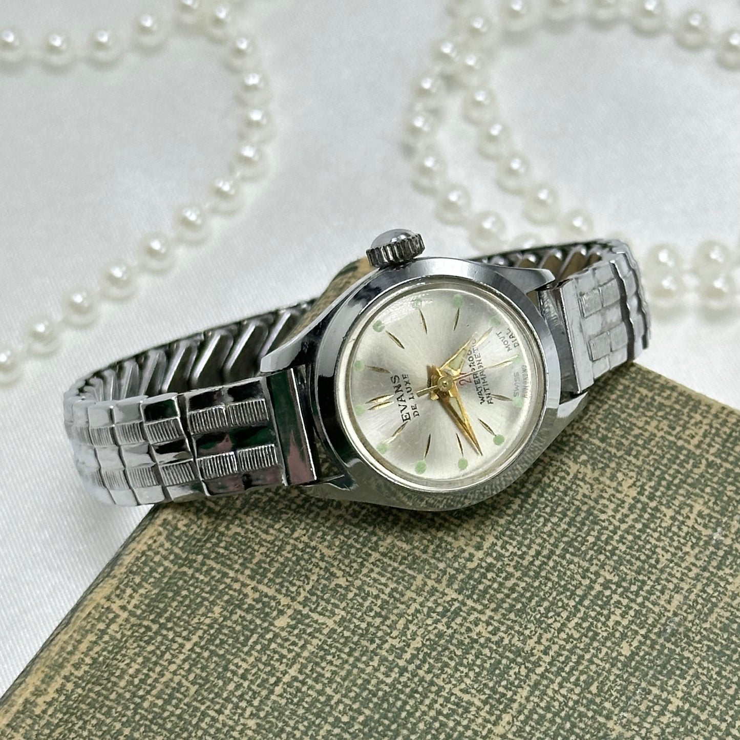 Vintage silver tone round dial Evans De Luxe 27 jewel manual winding Swiss made watch with gold hands and green markers. Band is stretchable. Waterproof and antimagnetic.