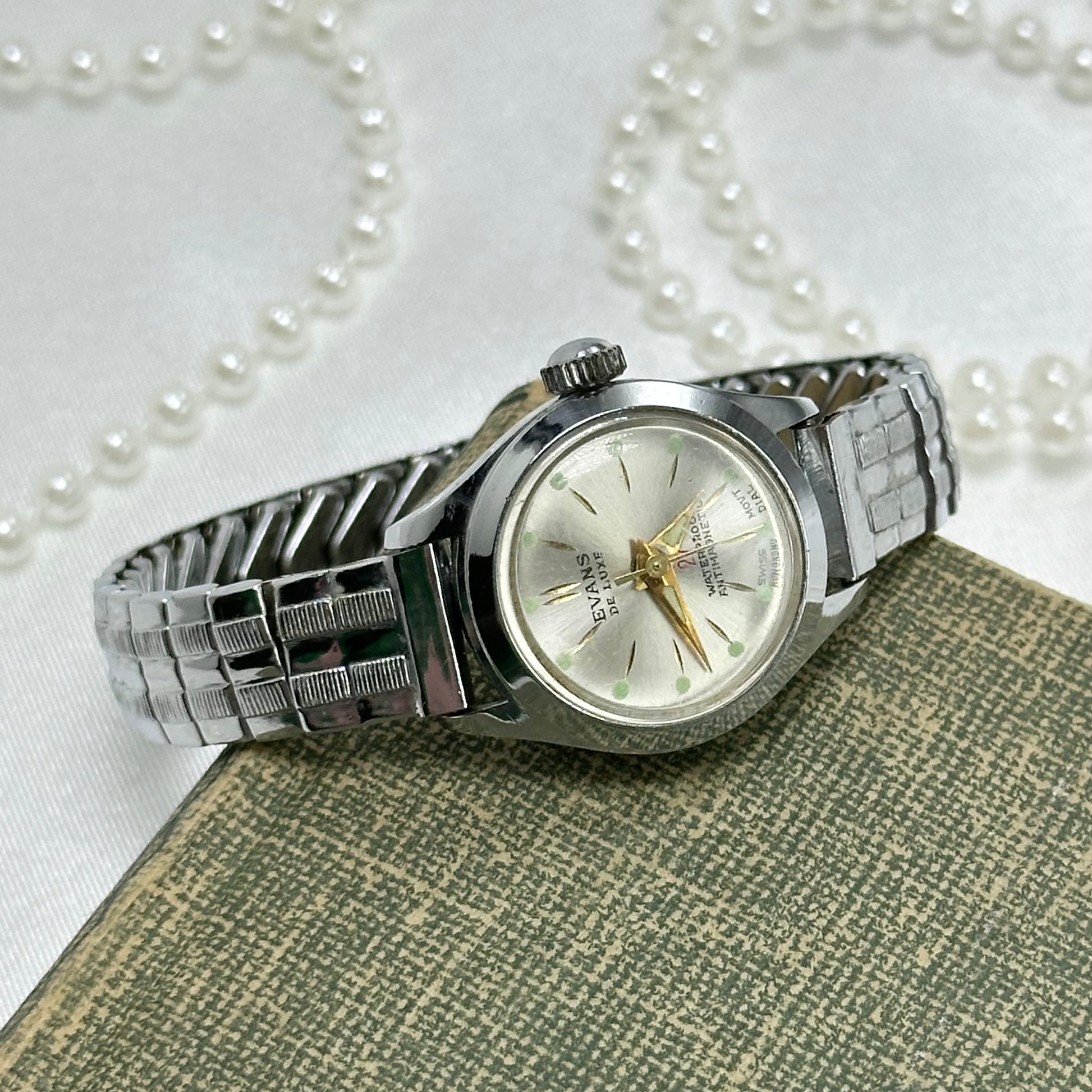Vintage silver tone round dial Evans De Luxe 27 jewel manual winding Swiss made watch with gold hands and green markers. Band is stretchable. Waterproof and antimagnetic.
